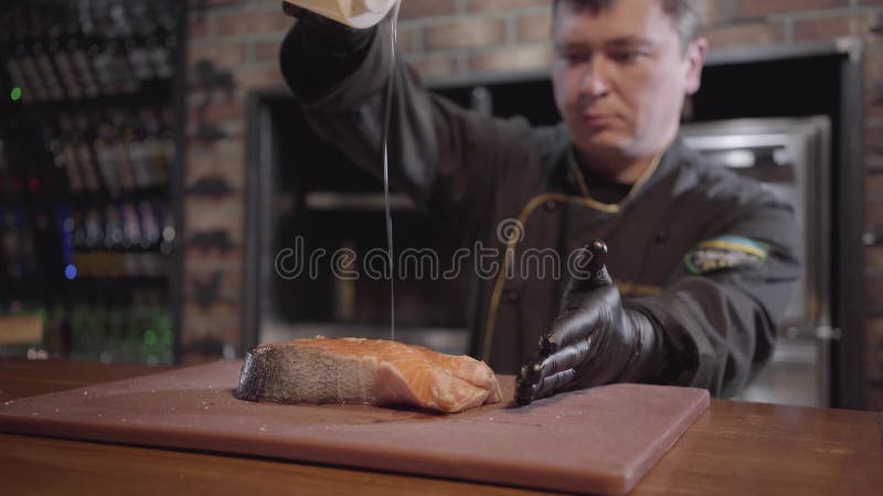Шеф-повар портрета полить большую часть семг с концом масла вверх Человек в черной робе и резиновых перчатках варя блюдо подготов